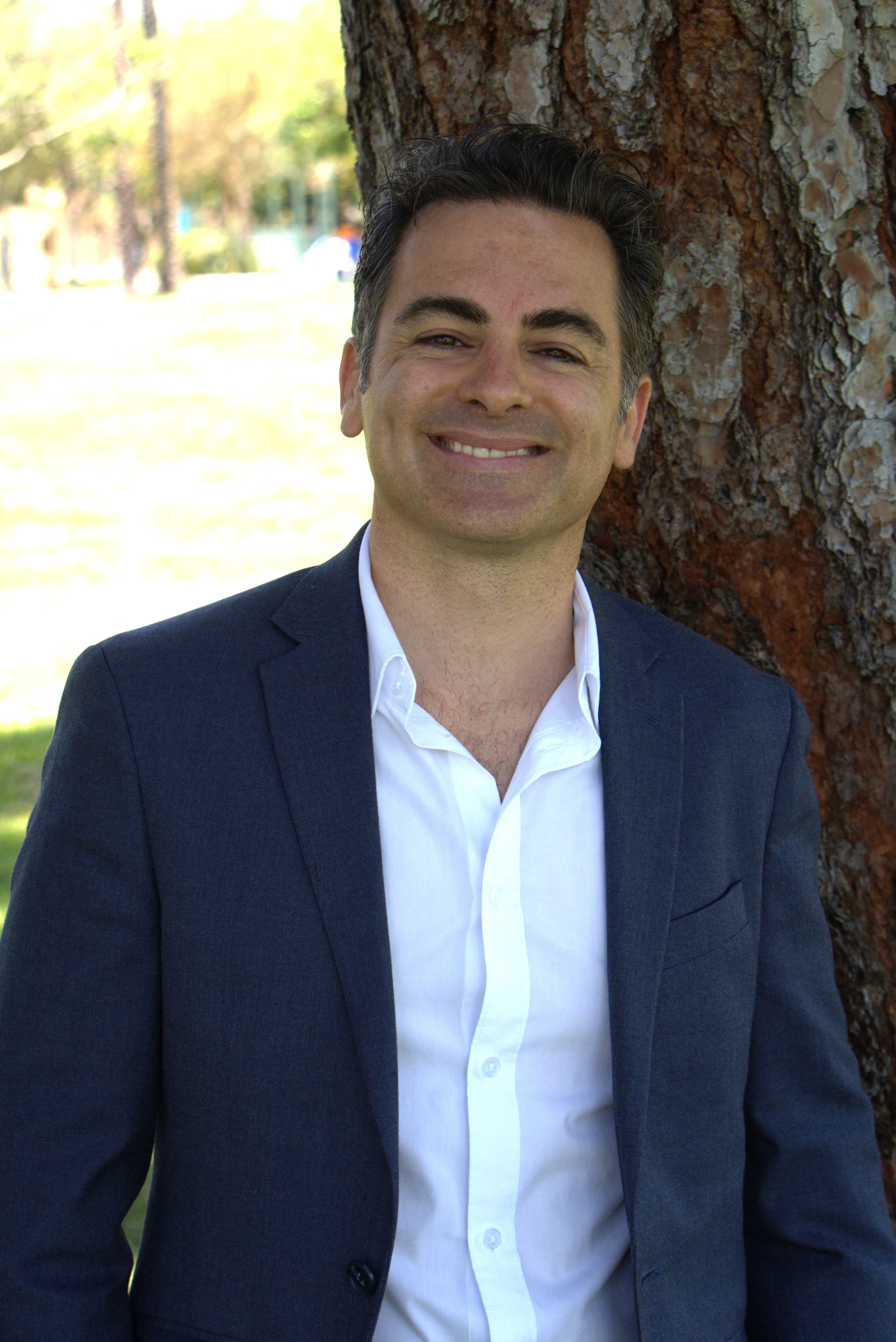 Smiling man in a navy blazer and white shirt leaning against a tree outdoors.